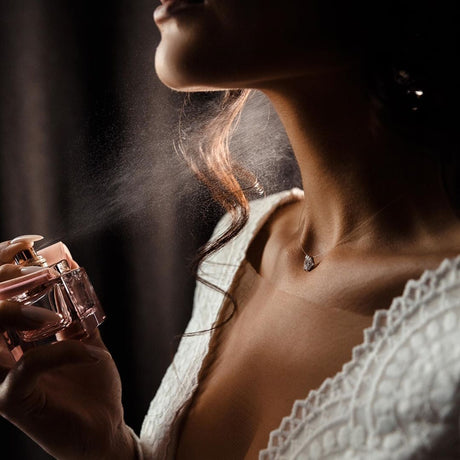 alt="Woman in white lace sprays pink perfume, mist suspended mid-air, with chain jewelry at her collarbone, against amber backdrop"