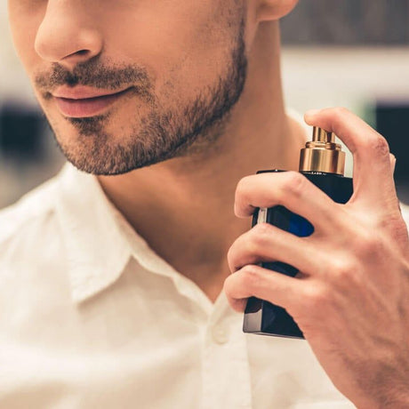 alt="Man with groomed beard holds black perfume bottle with gold pump, white shirt cuff visible, against blurred ivory backdrop"