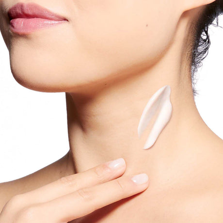 alt="Woman’s neck with contoured jawline, applying white moisturizing cream diagonally across her throat, with pink lips and manicured nude nails, against a white backdrop"