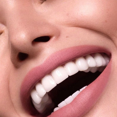 alt="Woman's smiling mouth with pale pink lips and white teeth, nostrils framing the composition, against blurred skin tone background"