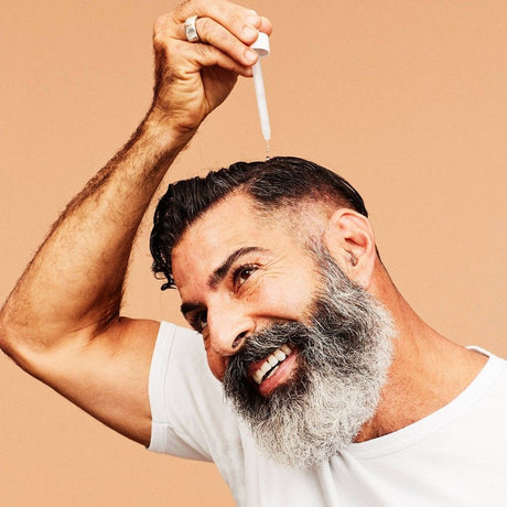alt="Man with salt-and-pepper beard applies liquid from white dropper bottle to black hair, against beige backdrop"