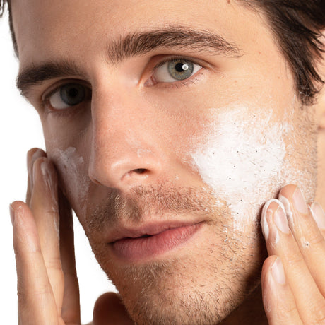 alt="Man with teal-blue eyes and stubbled jawline, hands framing his face with white foam, mid-cleansing, against a white backdrop"
