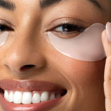 alt="Woman applying pale pink hydrogel eye patches under her eyes in a close-up, with soft pink lips and natural skin tone"