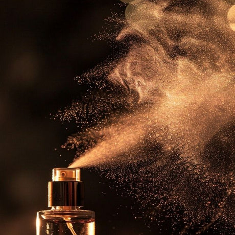 alt="Gold metal perfume bottle spraying fine mist upwards with droplets glittering on black background"