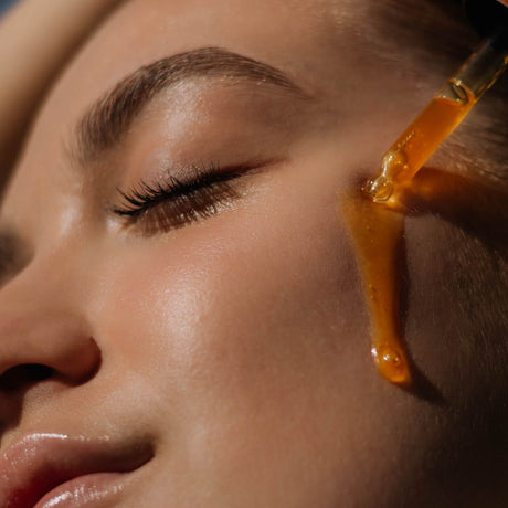 alt="Woman with closed eyes and contoured brows receiving serum from a pipette near her glowing cheek, against a blurred backdrop"