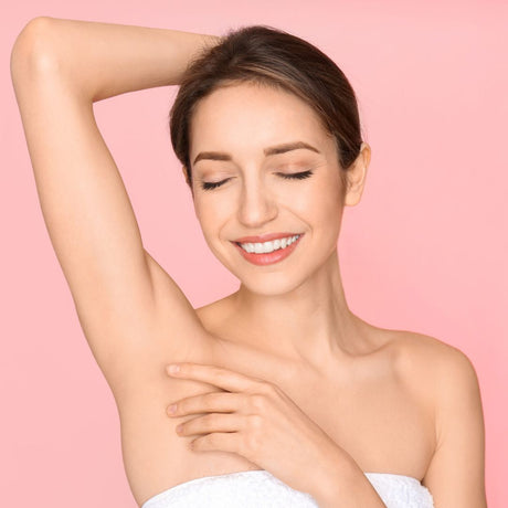 alt="Smiling woman wrapped in a white towel, resting her hand on her head and cradling her torso against a soft pink backdrop"