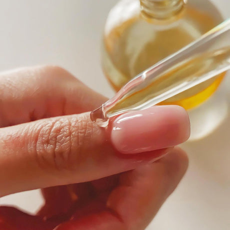alt="Manicured hand with pink nails receiving serum from a pipette, above a blurred amber skincare bottle"