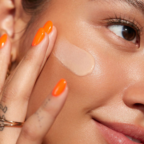 alt="Woman with brown eyes and voluminous lashes, her orange nails gently touching beige foundation on her cheek, against a solid backdrop"