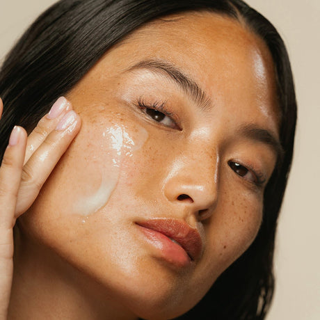 alt="Woman with black hair and freckled skin applying cream to her cheek, gazing aside against a neutral backdrop"