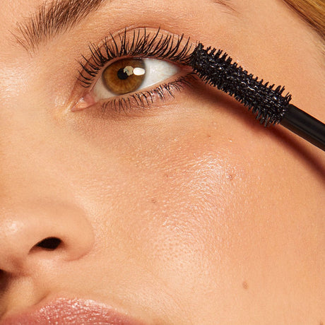 alt="Woman’s eye with long lashes being coated by black mascara, with glowing brown iris and natural skin tone, against a neutral backdrop"