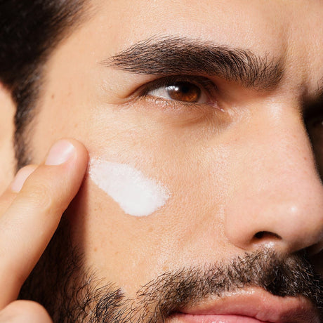 alt="A bearded man applying pearlescent cream to his cheekbone, with warm light accentuating his jawline and skin texture."