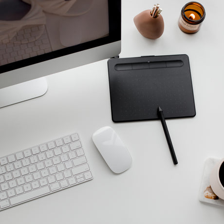 alt="Minimalist desk setup with silver-edged monitor, white keyboard, black drawing tablet, tan figurine, and amber candle cup"