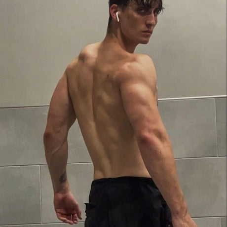alt="Muscular male figure with defined traps and delts, intricate arm tattoo, wireless earbud, and black trousers against cool-toned tile backdrop"