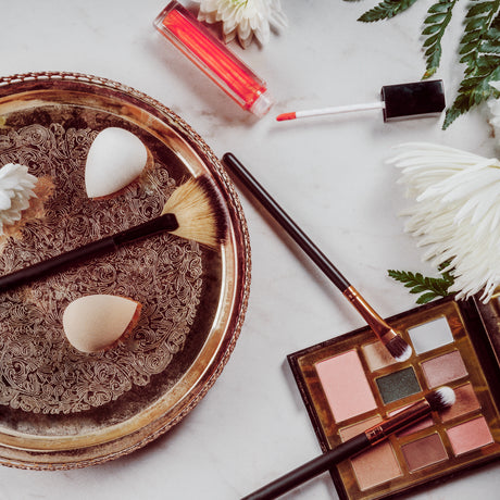 alt="A makeup set on a golden tray: a beauty sponge, brushes, eyeshadow palette and lip gloss"