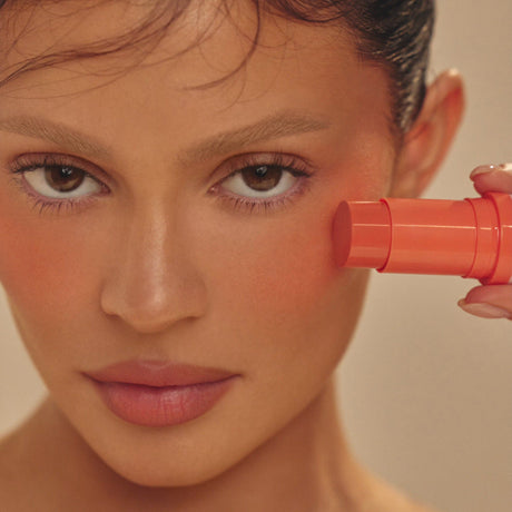 alt="Woman applying orange-red cosmetic stick to her flushed cheek, with natural makeup, against a light-toned backdrop"