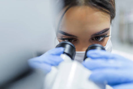 alt="Female researcher operating a microscope in a sterile lab environment, wearing mask and blue gloves"