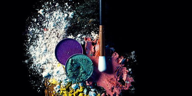 alt="Two eyeshadow palettes in purple and teal, with a makeup brush and colorful pigment dust on a black background"